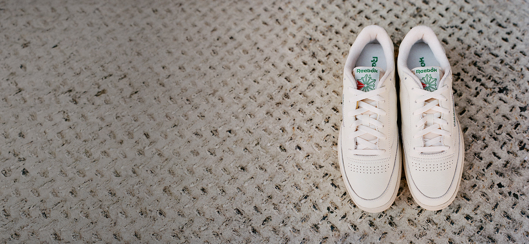 A pair of white Reebok sneakers with green and red logo details is placed neatly on a textured, light-colored carpet, positioned on the right side of the image.
