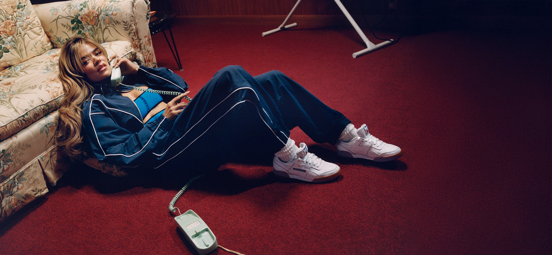 Person sitting on a red carpet with a phone and remote control, wearing blue tracksuit and white sneakers.