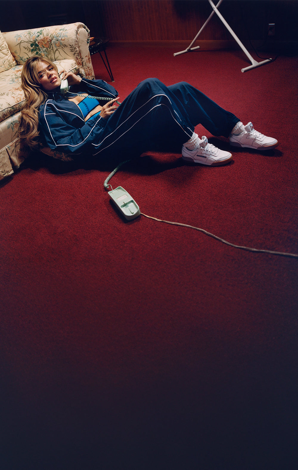 Person sitting on a red carpet with a phone and remote control, wearing a blue tracksuit.