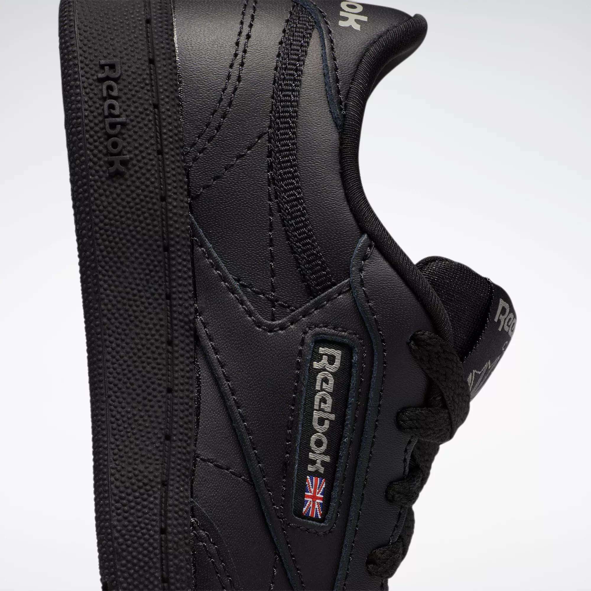 Close-up of the Reebok Kids' Club C Shoes for little kids, featuring a black leather upper with the Reebok logo and a small British flag patch on the side. The shoe is slightly tilted to show its side profile and textured sole.