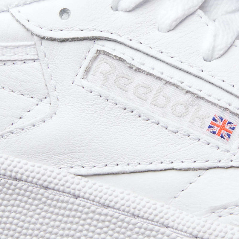 Close-up of Reebok Women's Club C 85 Shoes featuring an embossed Reebok logo, a small Union Jack flag on the side, and visible leather texture with detailed stitching.