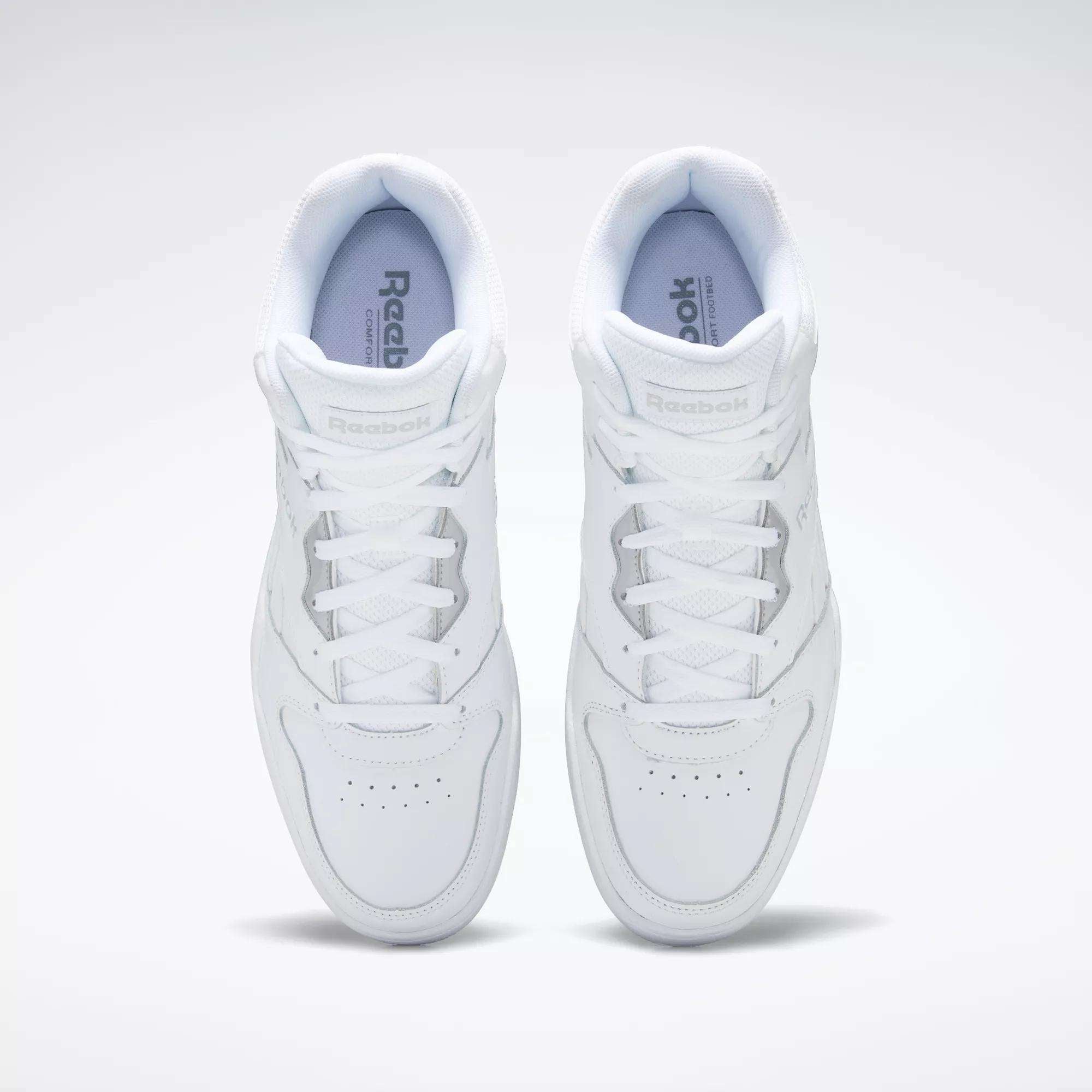 A pair of white Reebok Men's Royal BB4500 Hi 2.0 shoes viewed from above, highlighting the laces, perforated toe area, and Reebok branding on the tongues and insoles against a light, '80s-inspired background.