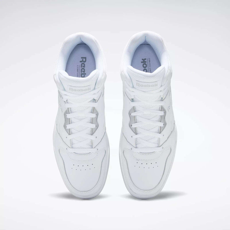 A pair of white Reebok Men's Royal BB4500 Hi 2.0 shoes viewed from above, highlighting the laces, perforated toe area, and Reebok branding on the tongues and insoles against a light, '80s-inspired background.