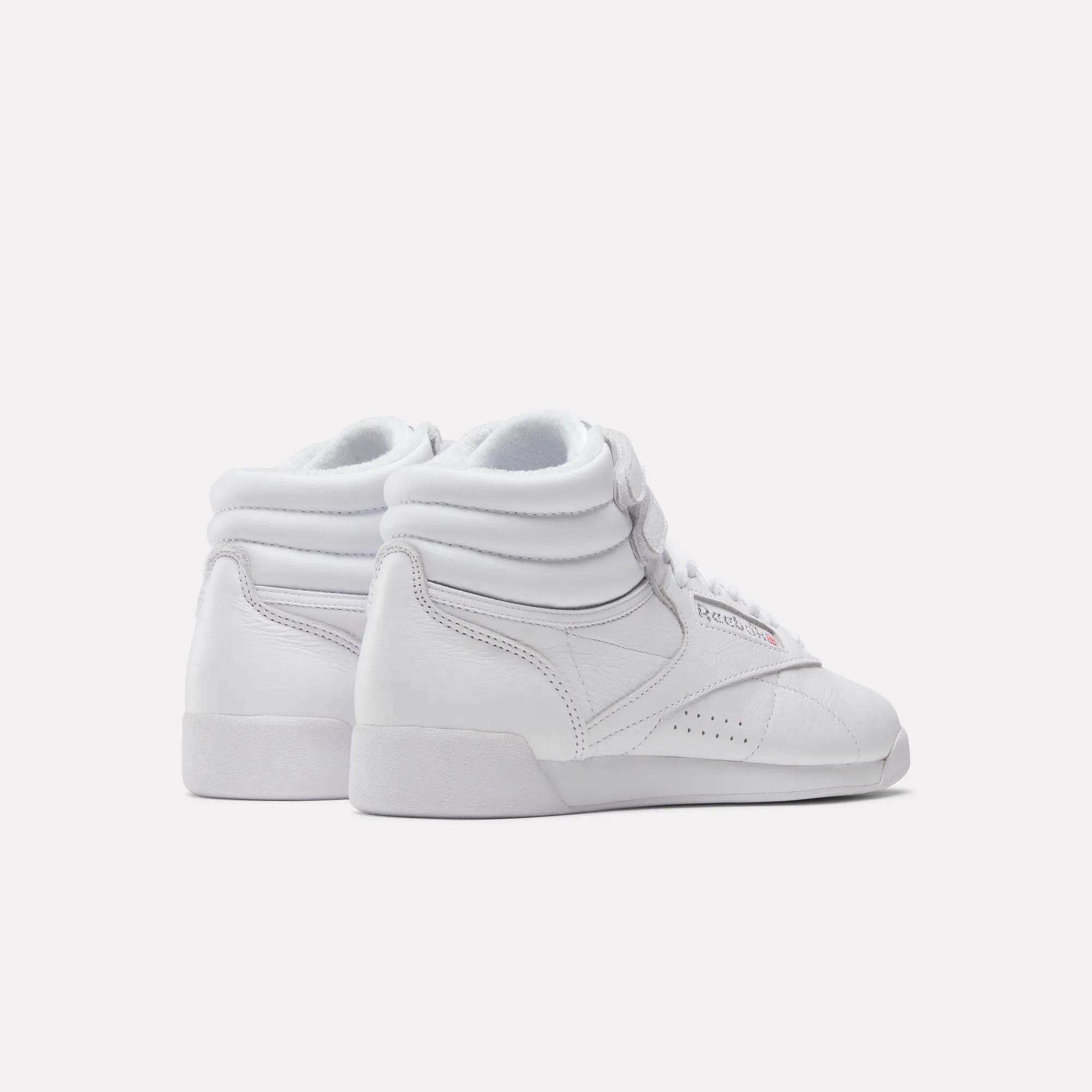 A pair of Reebok Women's Freestyle Hi Shoes in white, featuring padded ankles, side stitching, and white laces, displayed on a light background and angled away.