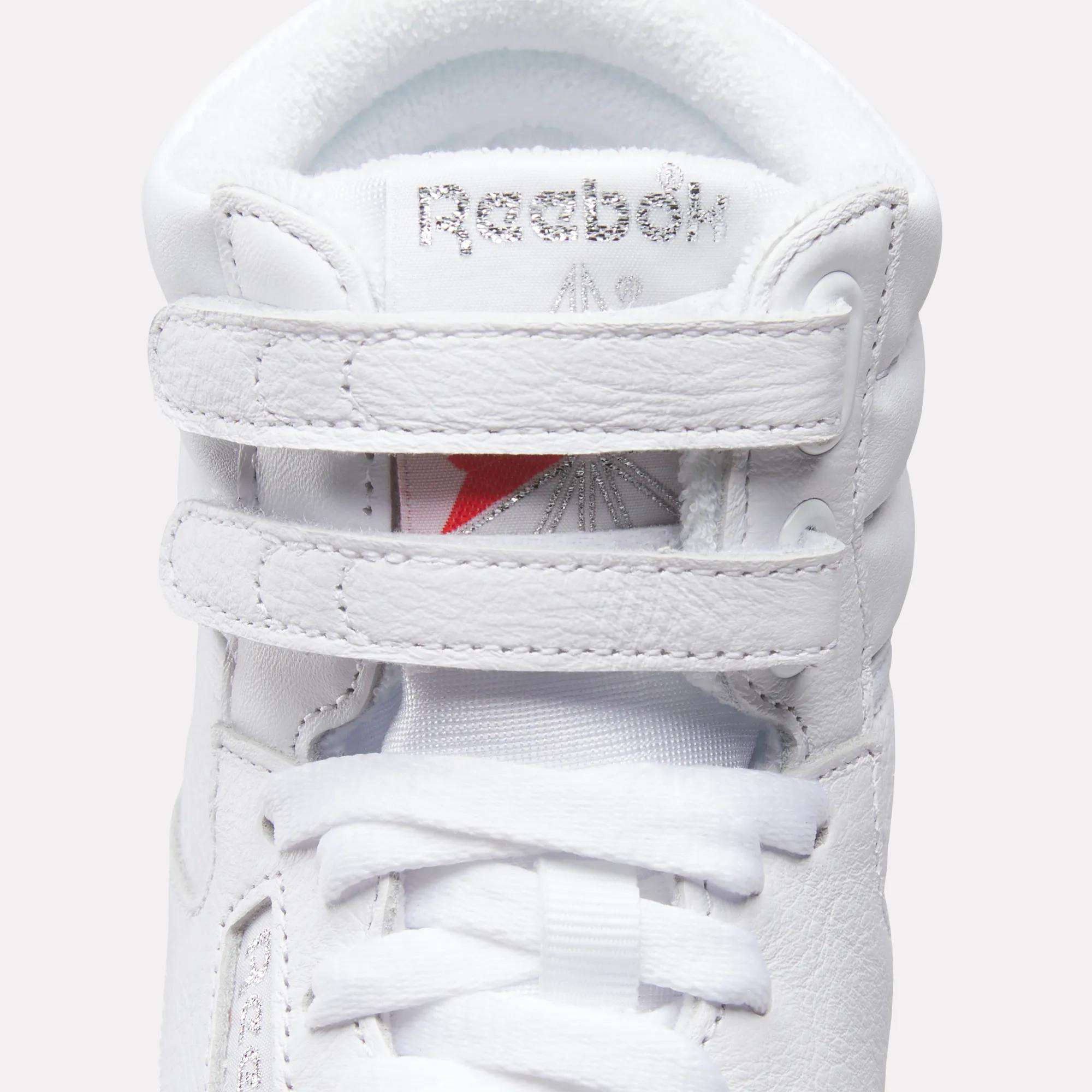 A close-up of the Reebok Women's Freestyle Hi Shoes, featuring a white textured leather design with two velcro straps, white laces, and a silver Reebok logo on the tongue—ideal for women's fitness.