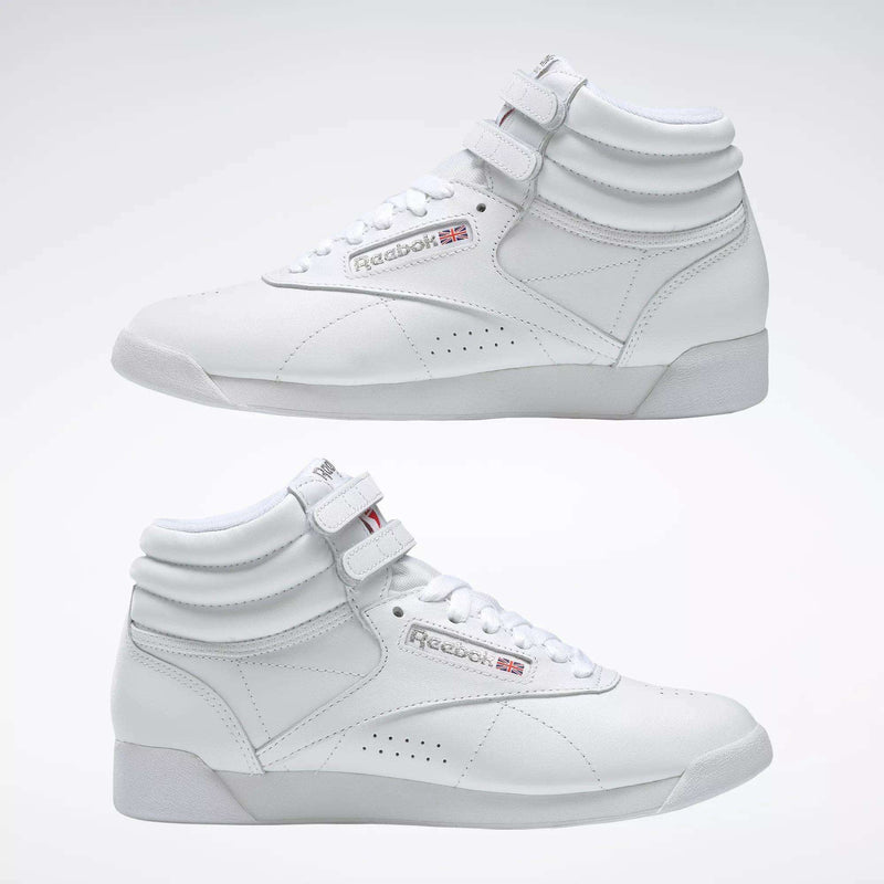 Two Reebok Women's Freestyle Hi Shoes in white are shown side by side on a light background, highlighting both outer and inner sides. Features include visible laces, velcro straps, and classic Reebok logos—ideal for women's fitness.