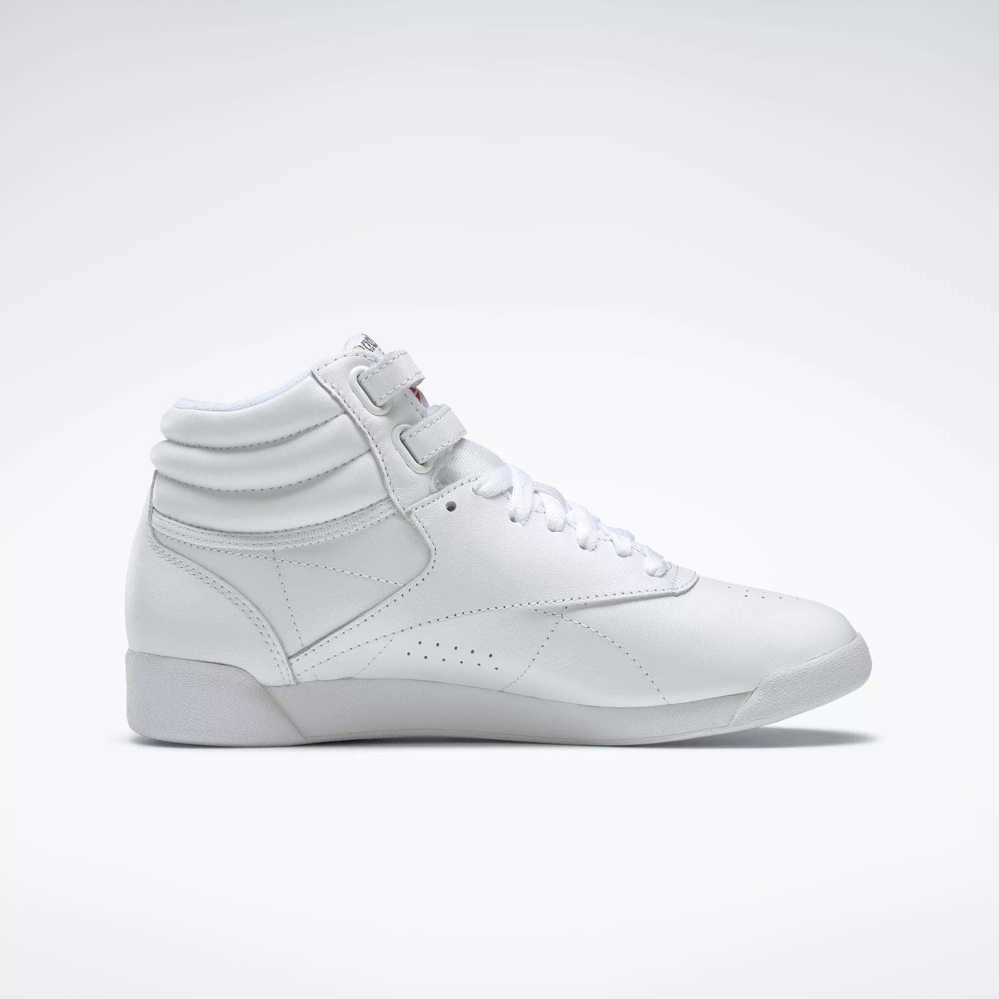 The Reebok Women's Freestyle Hi Shoes are white high-top sneakers with laces and velcro straps, showcasing a minimalist design and subtle stitching—perfect for women's fitness routines. Shown in a side view on a plain light background.