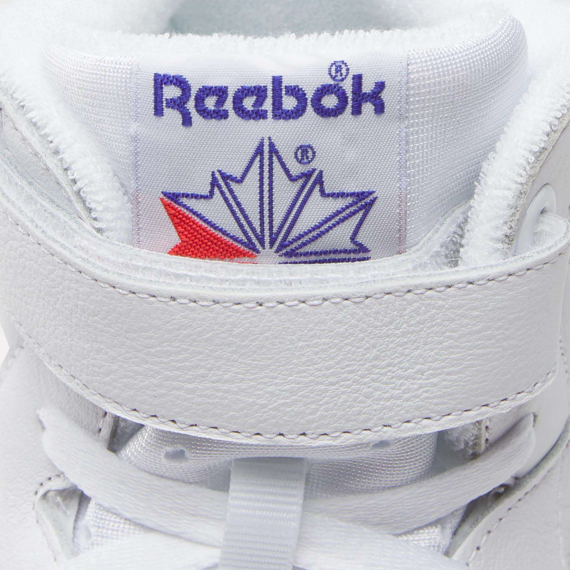 Close-up of Reebok Men's EX-O-FIT Hi Shoes in white, featuring a high-cut design, blue Reebok logo on the tongue, red and blue star detail, white laces, and a retro velcro strap.