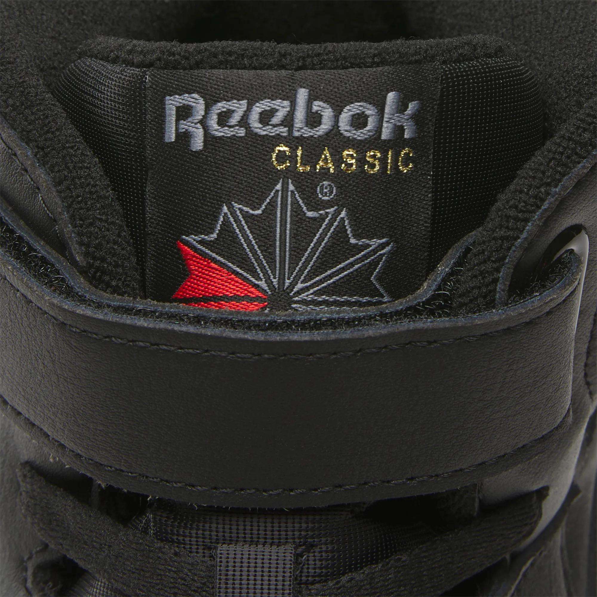 Close-up of a black Men's EX-O-FIT Hi Reebok shoe showing the tongue with the Reebok logo, CLASSIC in gold, and a red and gray stylized maple leaf, highlighting its retro look.