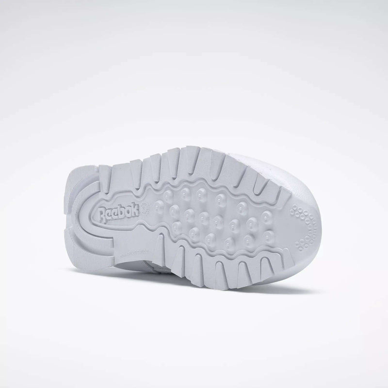 The image displays the sole of a Kids' Classic Leather Shoe - Preschool by Reebok lying on its side, showcasing the textured grip, lightweight cushioning, and the embossed Reebok logo.