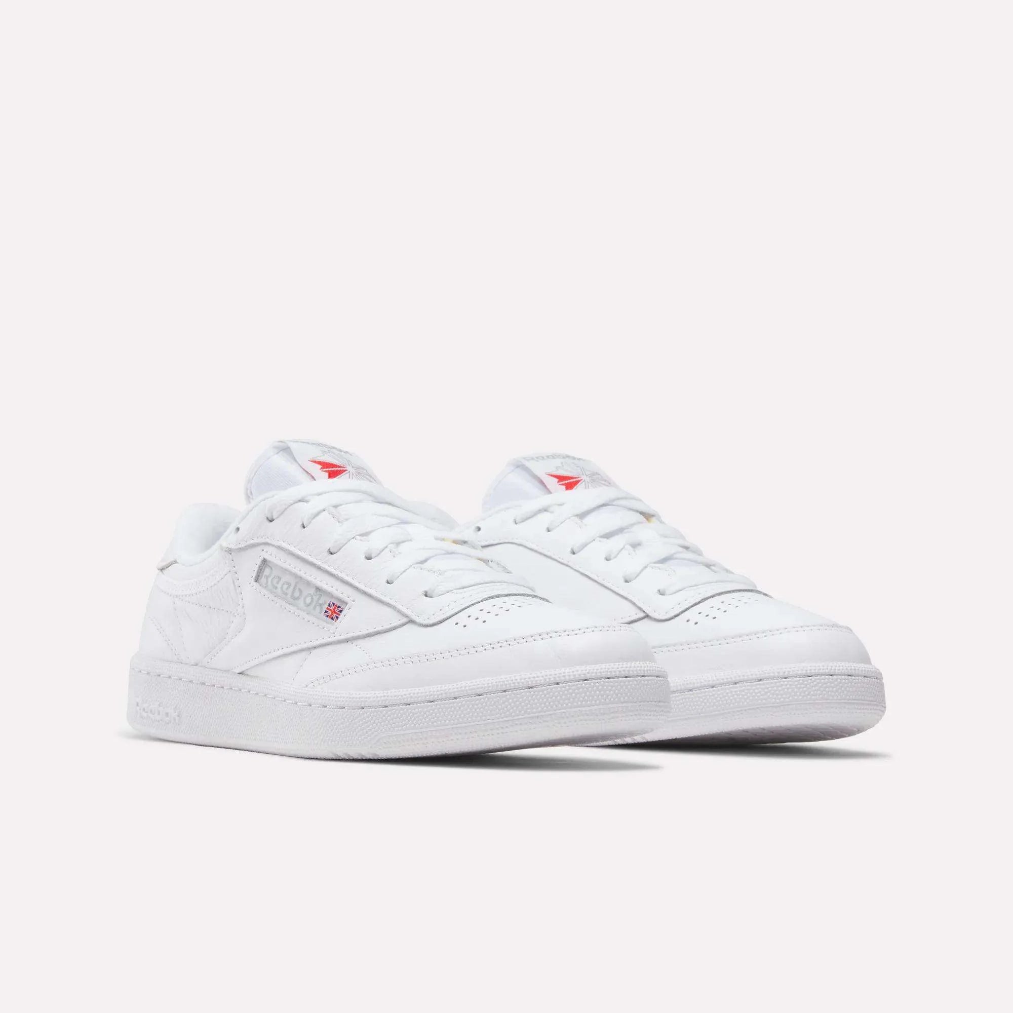 A pair of white Reebok sneakers with low tops, featuring the Reebok logo and a small red and blue flag on the side, displayed on a light background.