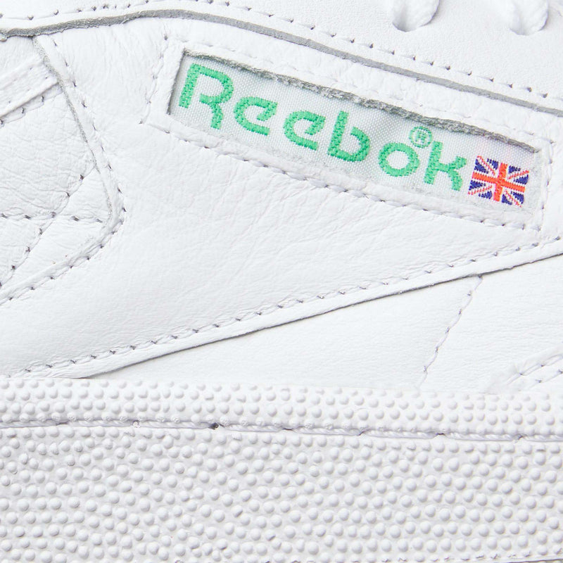 Close-up of Reebok Club C 85 Shoes in white leather, featuring a green Reebok logo and Union Jack flag on the side label, detailed stitching, and a textured rubber sole that showcases its tennis-inspired design.