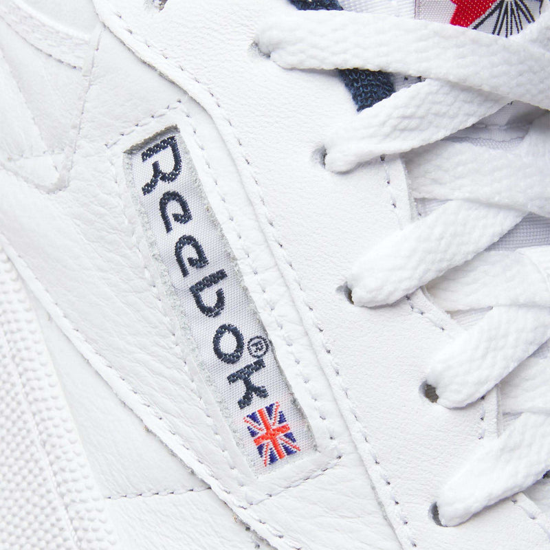 Close-up of the Reebok Club C 85 Shoes in white leather, featuring textured uppers, white laces, and a side label with the Reebok logo and Union Jack flag. These tennis-inspired sneakers blend iconic style with quality craftsmanship.