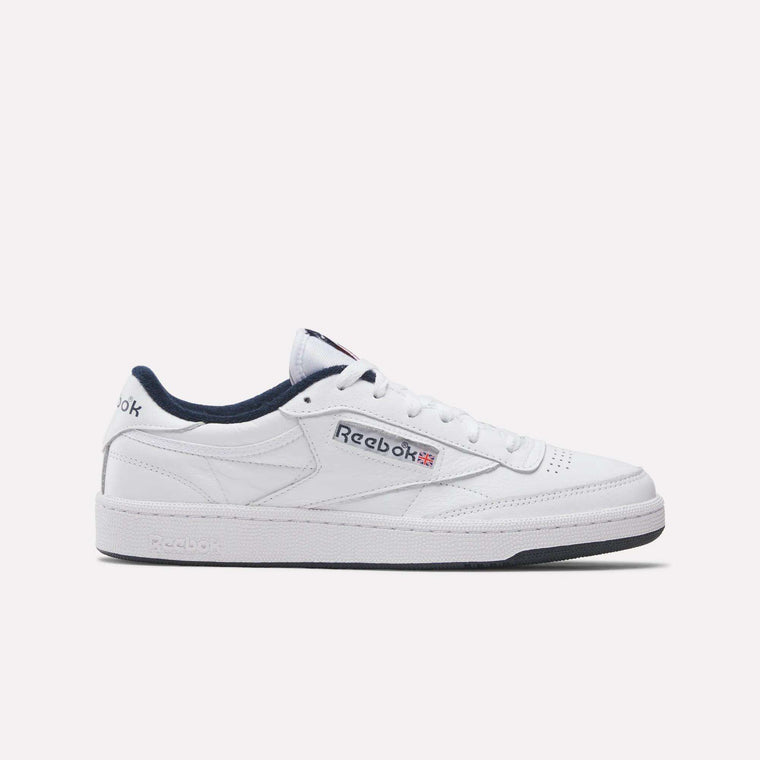 A white Reebok Club C 85 shoe with navy blue details, the Reebok logo, and a Union Jack flag on the side sits on a plain background. These classic tennis-inspired leather sneakers by Reebok feature timeless style.