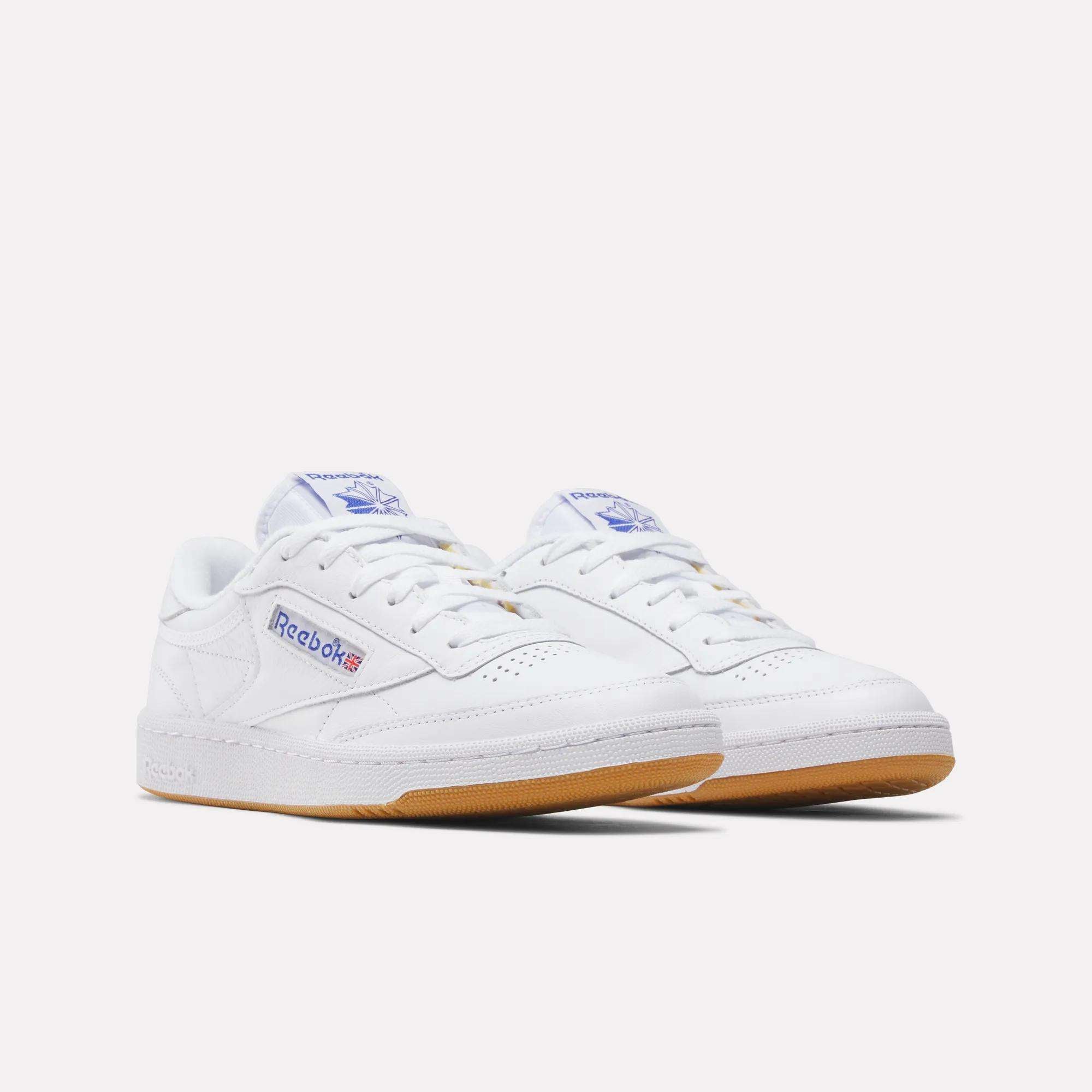 A pair of Reebok Club C 85 Shoes in white with gum soles, blue Reebok logos on the sides and tongues, plus subtle red accents, displayed against a plain white background.