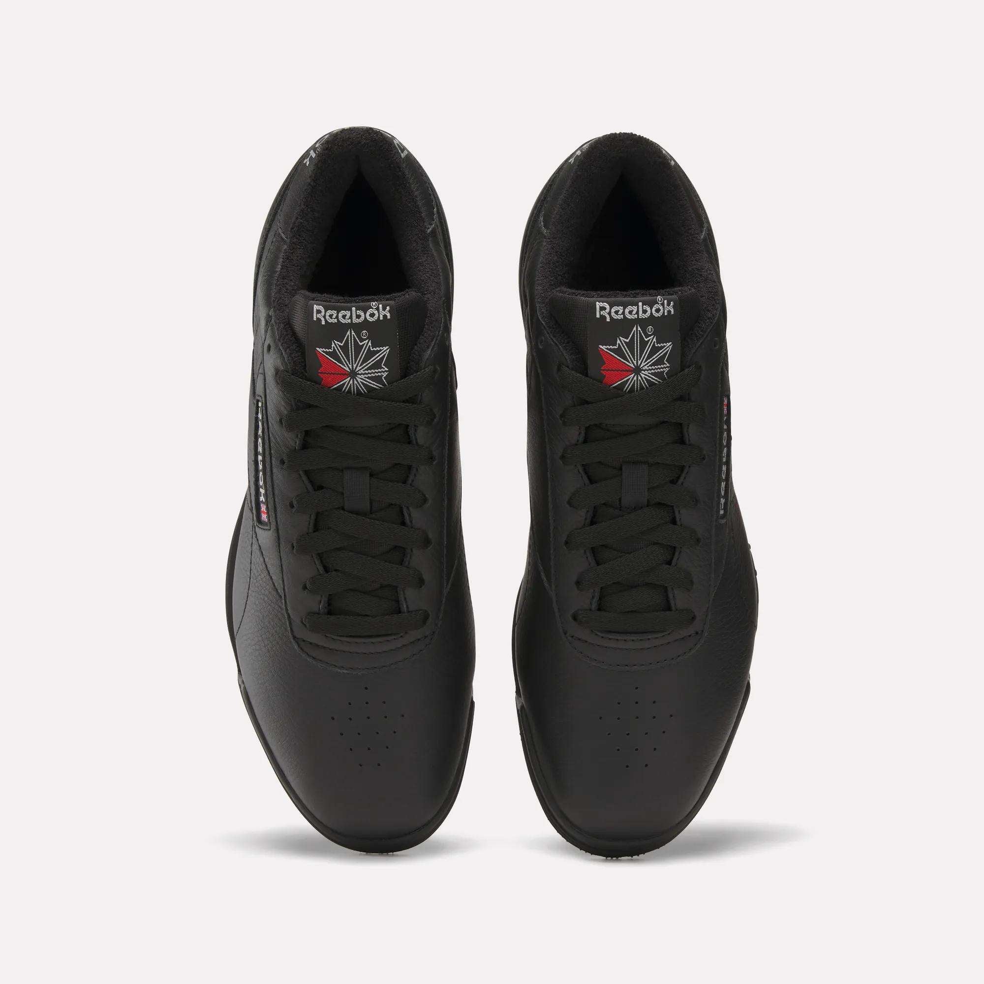 Top view of Reebok Men's Exofit Lo Shoes in black with black laces, the Reebok logo, and a red and white starburst on each tongue. The low-profile shoes are placed side by side on a light background.