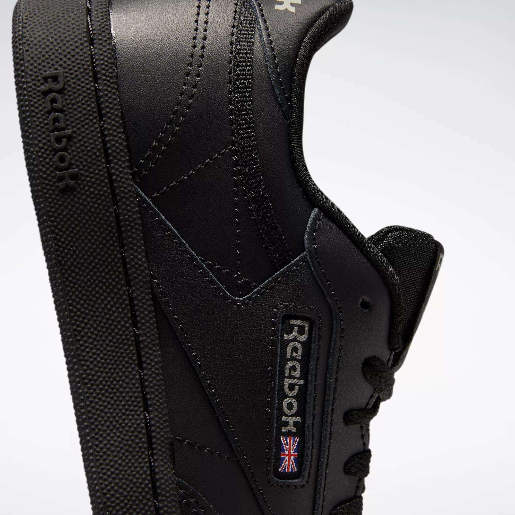 Close-up of Reebok Kids' Club C Shoes - Grade School in black, featuring a side view with visible stitching, soft leather uppers, textured sole, and Reebok branding with a small Union Jack flag.