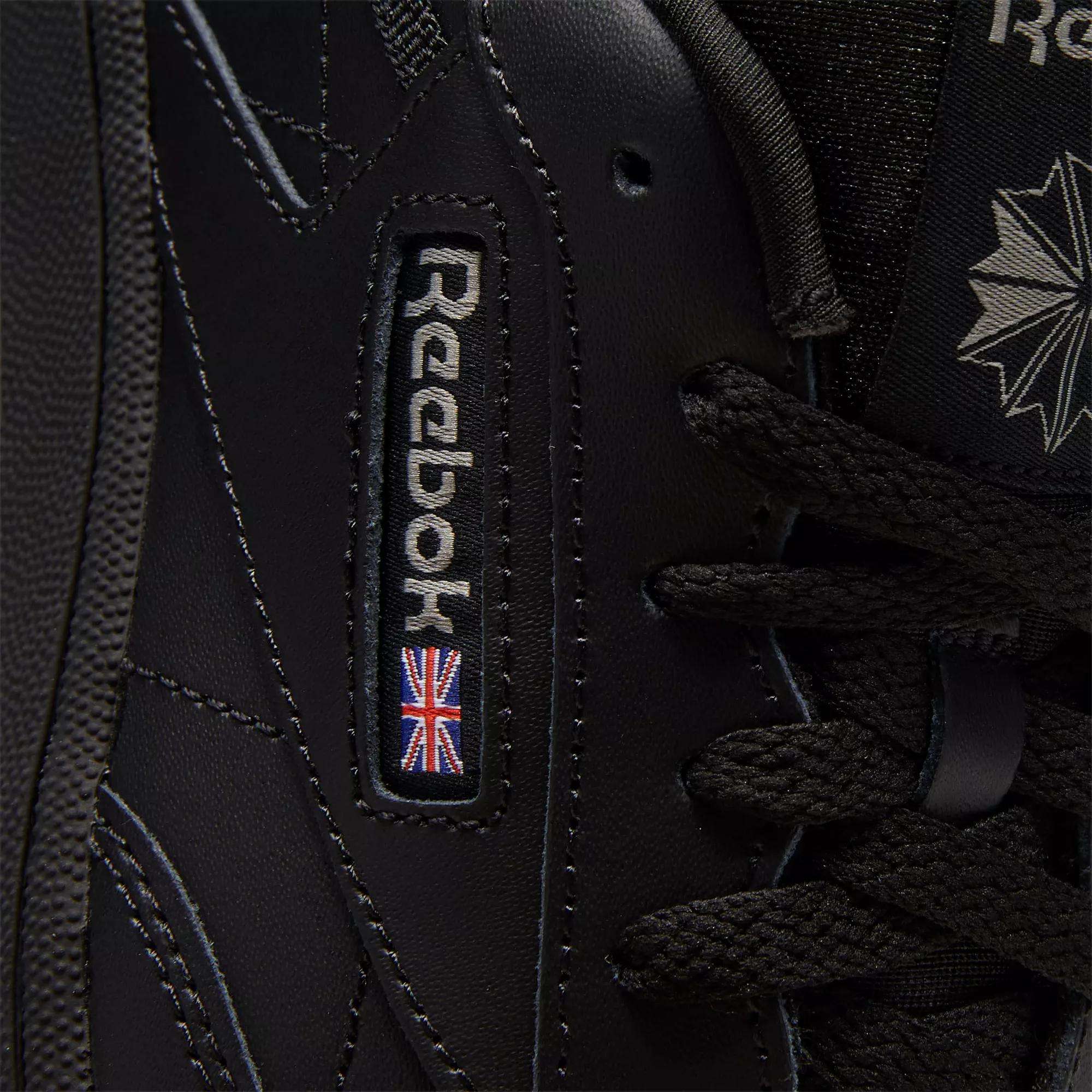 Close-up of black Reebok Kids' Club C Shoes - Grade School, showcasing classic court style with detailed stitching on soft leather uppers, plus the Reebok logo and a small UK flag patch near the laces.