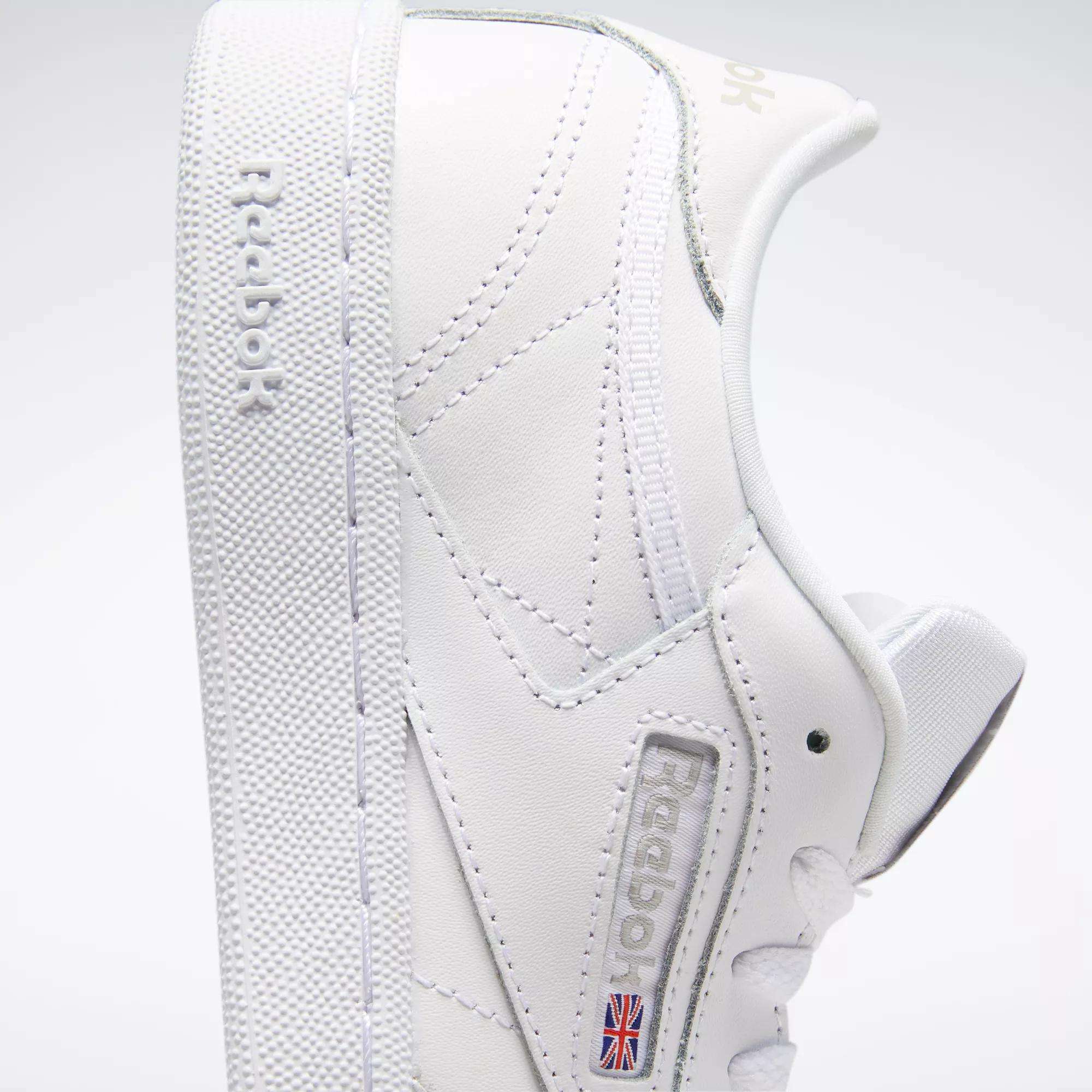 A close-up side view of the Reebok Kids' Club C Shoes - Grade School displays soft white leather uppers, clean minimal stitching, a textured sole, "Reebok" embossed on the side, and the Union Jack flag near the sole.