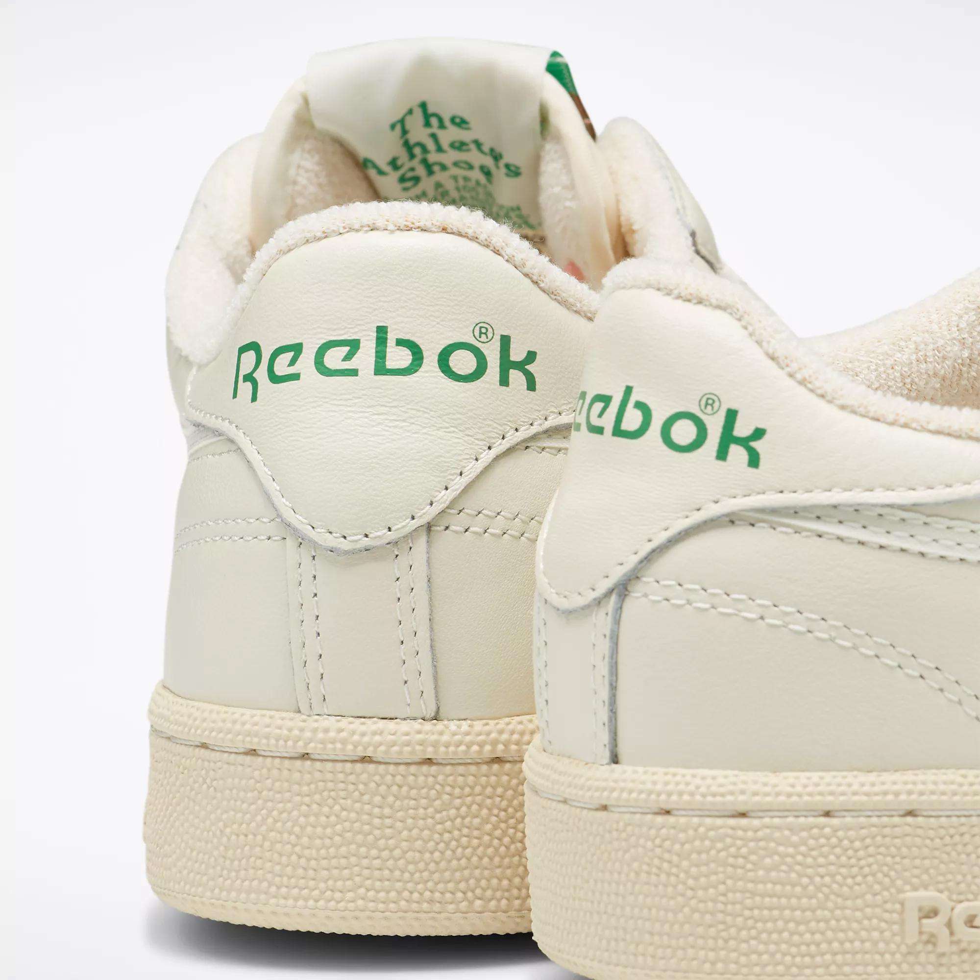 Close-up of the heels of two off-white Reebok Club C 85 Vintage Shoes with leather uppers, green Reebok logos, and textured rubber soles, set against a plain white background.