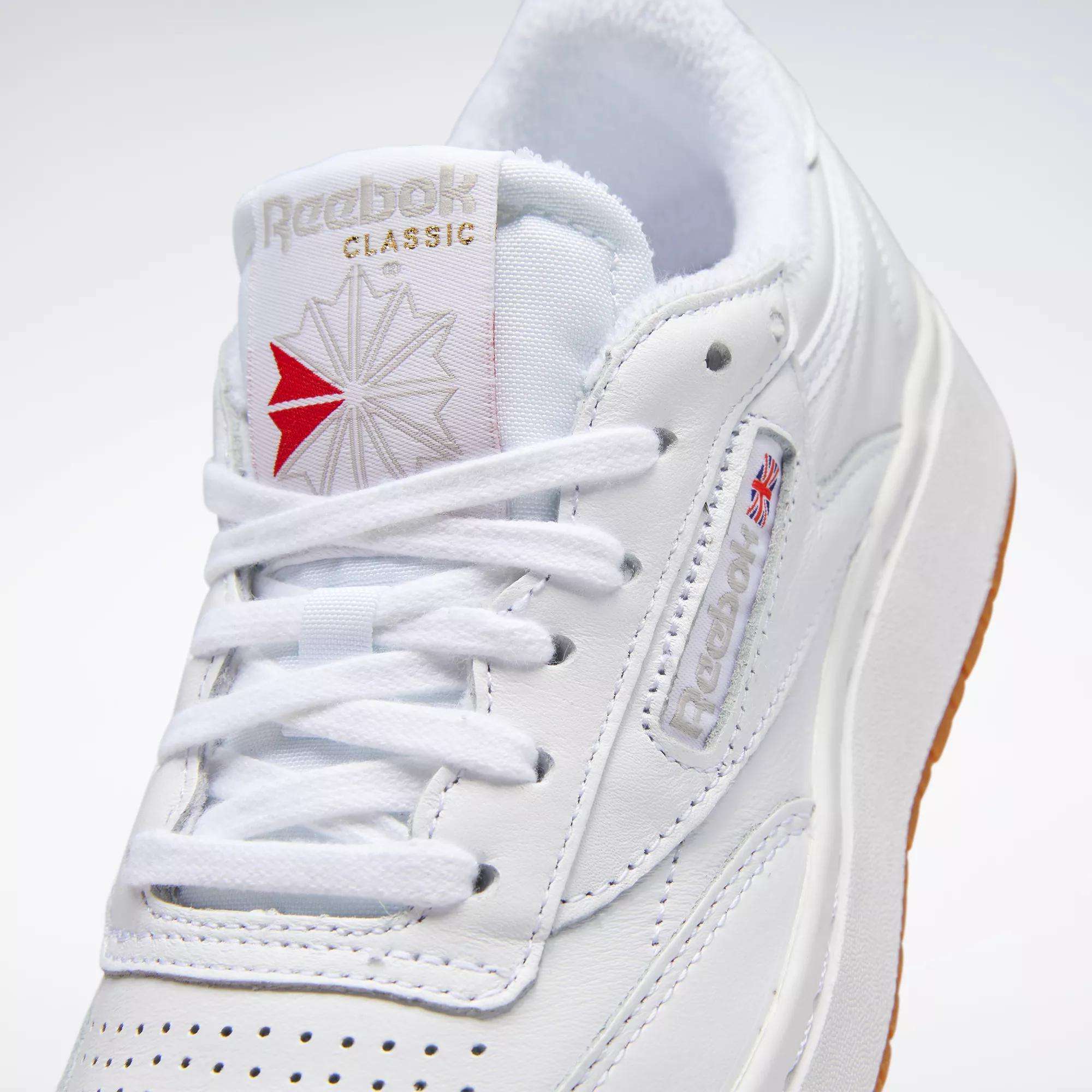 Close-up of the Reebok Women's Club C Double Shoes in white leather, featuring white laces, a red logo on the tongue, embossed side branding, a Union Jack flag detail, and a white sole with gum-colored bottom. Inspired by tennis style.
