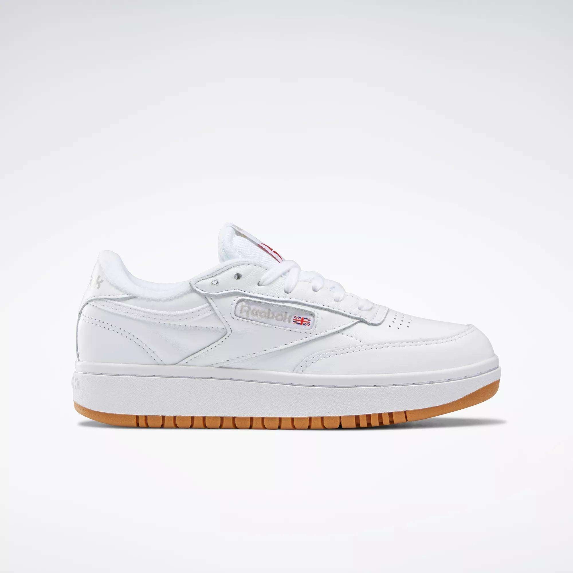 The Reebok Women's Club C Double Shoes are tennis-inspired white leather sneakers with a low profile, white laces, side stitching, gum rubber sole, and the Reebok logo with British flag on the side. Displayed on a plain light background.