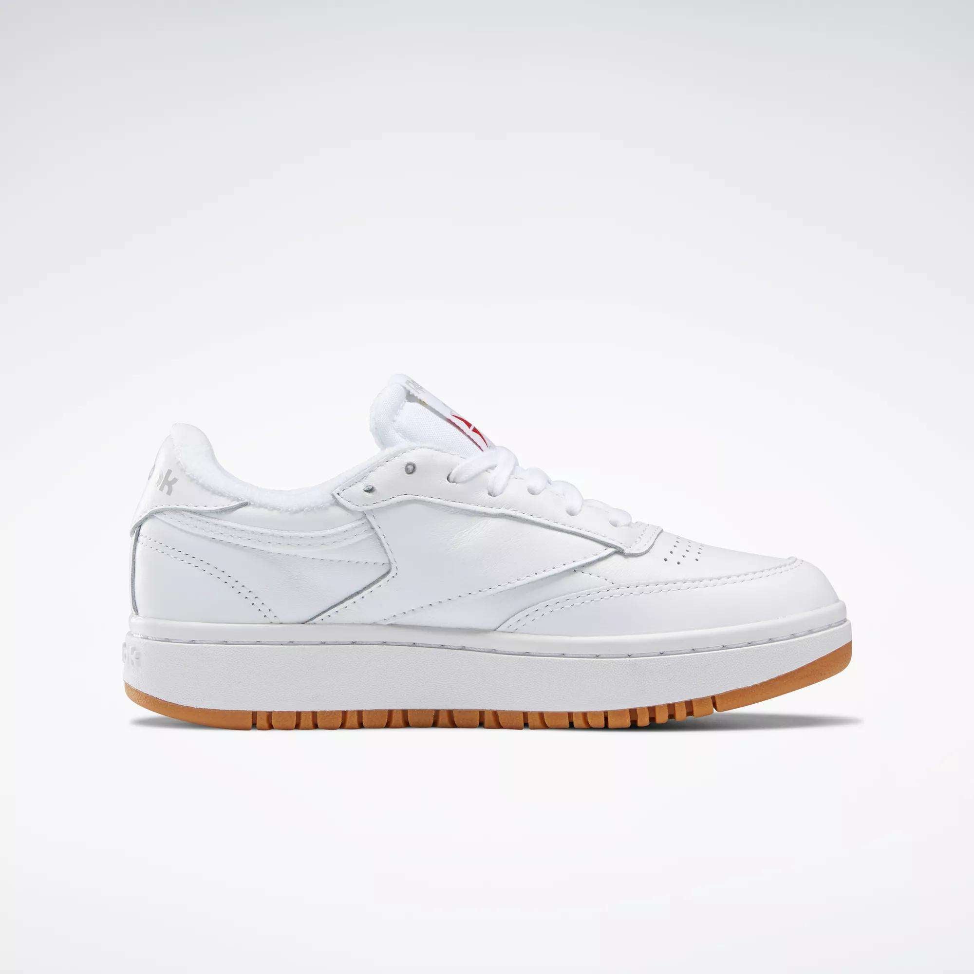 The Reebok Women's Club C Double Shoes are white, low-top sneakers with white laces, subtle stitching, and a light brown rubber sole with a platform midsole, shown in profile against a plain white background.
