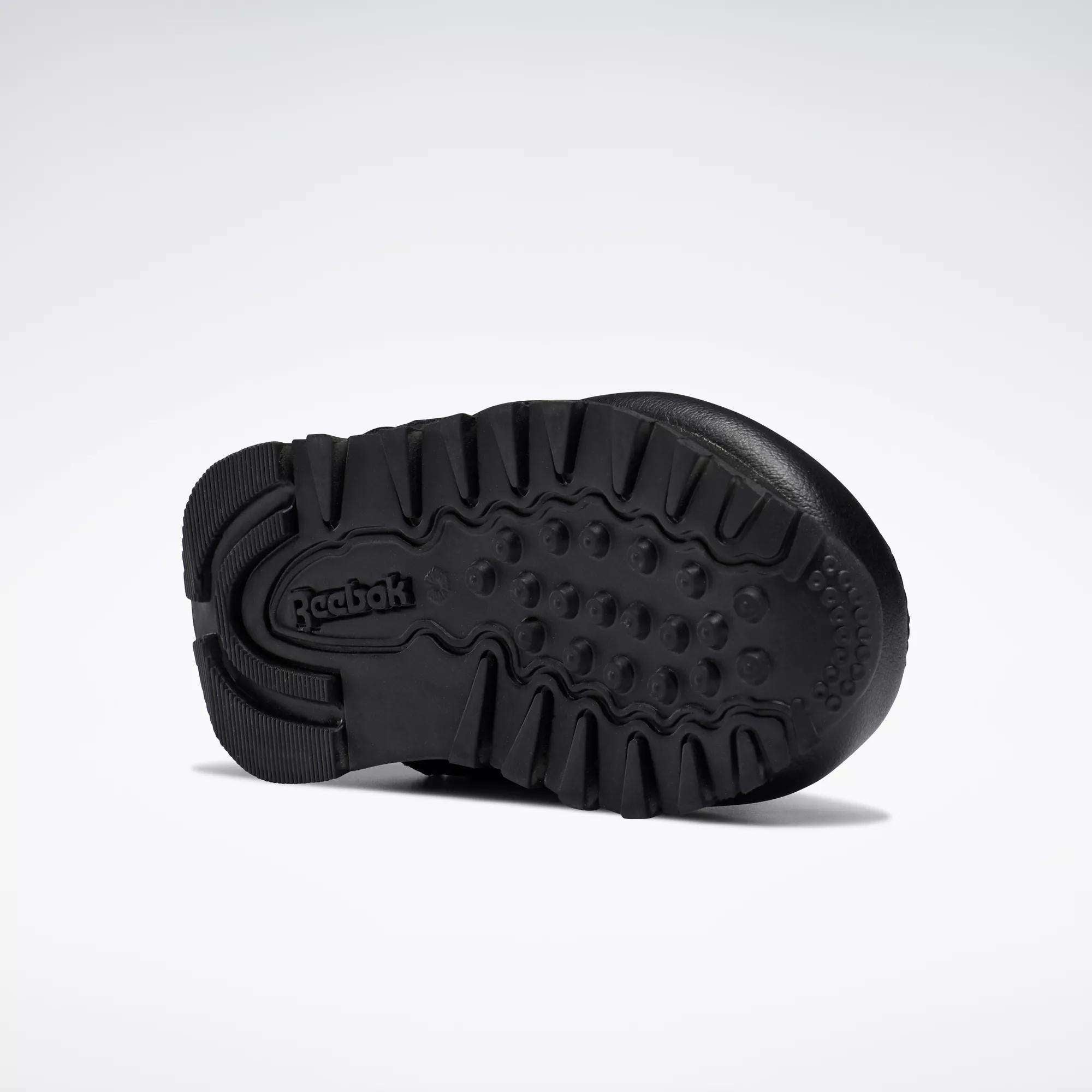 The image displays the black rubber sole of Reebok Kids' Classic Leather Shoes for babies and toddlers, featuring deep treads and the Reebok logo at the heel, set against a plain white background—ideal for young fans of classic sneakers.