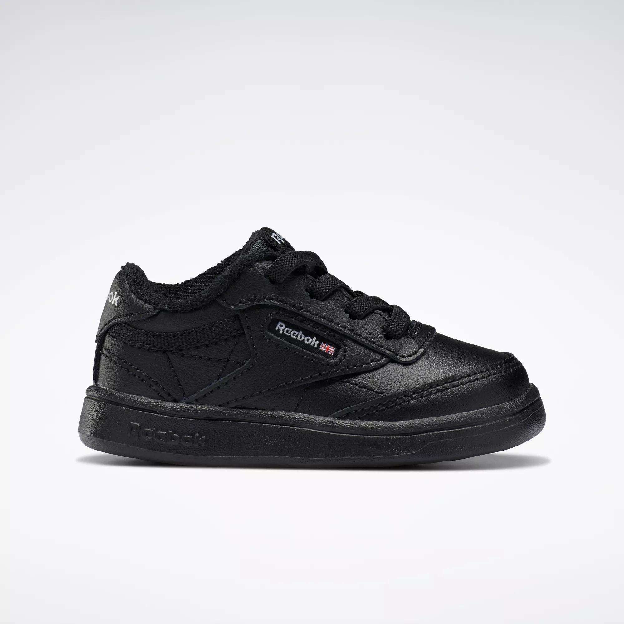 A side profile of the Reebok Kids' Club C Shoes for babies and toddlers is shown against a white background, featuring black soft leather, black laces, detailed stitching, and a small Reebok logo with British flag on the side.