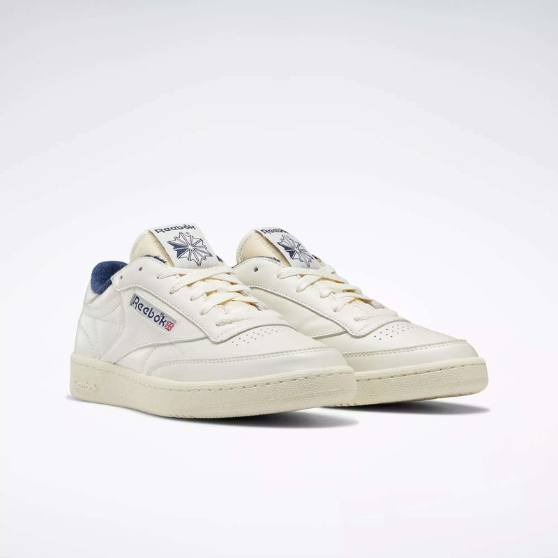 A pair of off-white Reebok Club C 85 Vintage Shoes with navy accents, featuring the Reebok logo and Union Jack on the side and tongue, along with lightweight cushioning for all-day comfort, set against a plain white background.