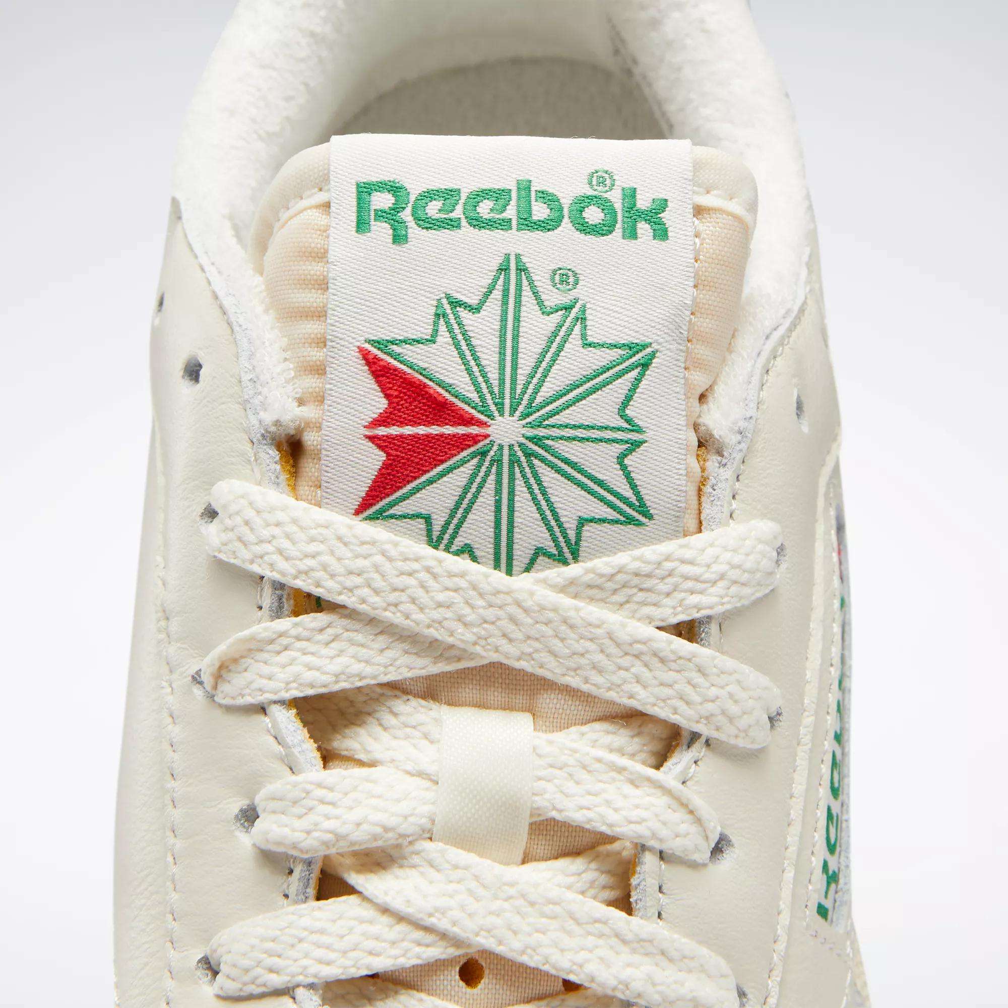 Close-up of Reebok Women's Club C 85 Vintage Shoes, featuring the green Reebok logo above a green and red starburst on the tongue, with cream laces and textured leather for a stylish, classic tennis look.