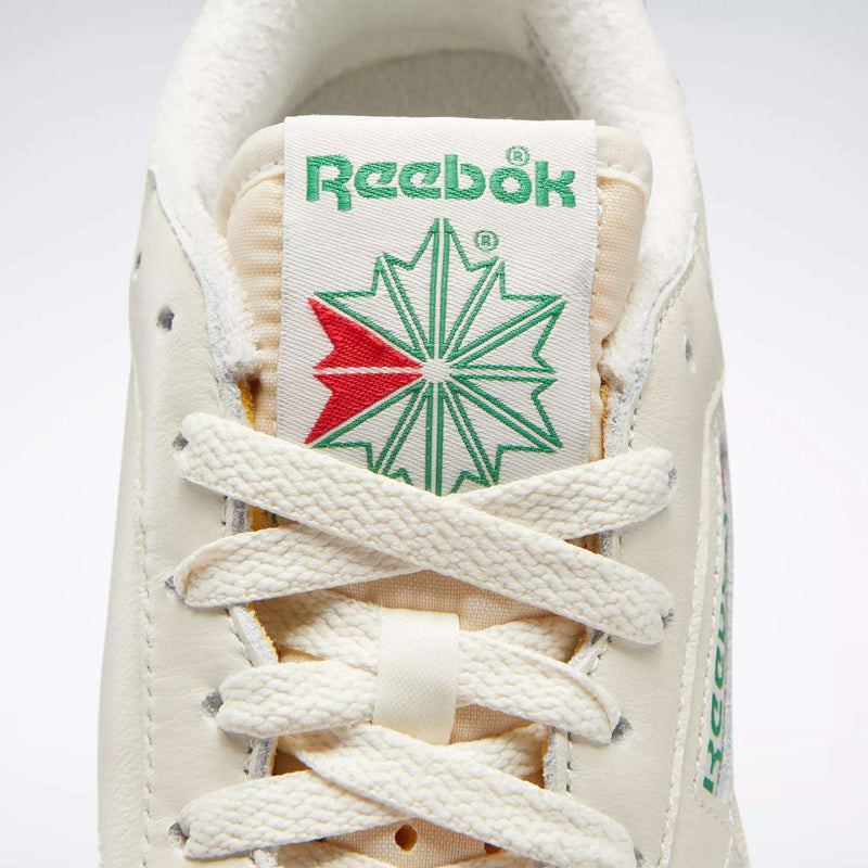 Close-up of Reebok Women's Club C 85 Vintage Shoes, featuring the green Reebok logo above a green and red starburst on the tongue, with cream laces and textured leather for a stylish, classic tennis look.