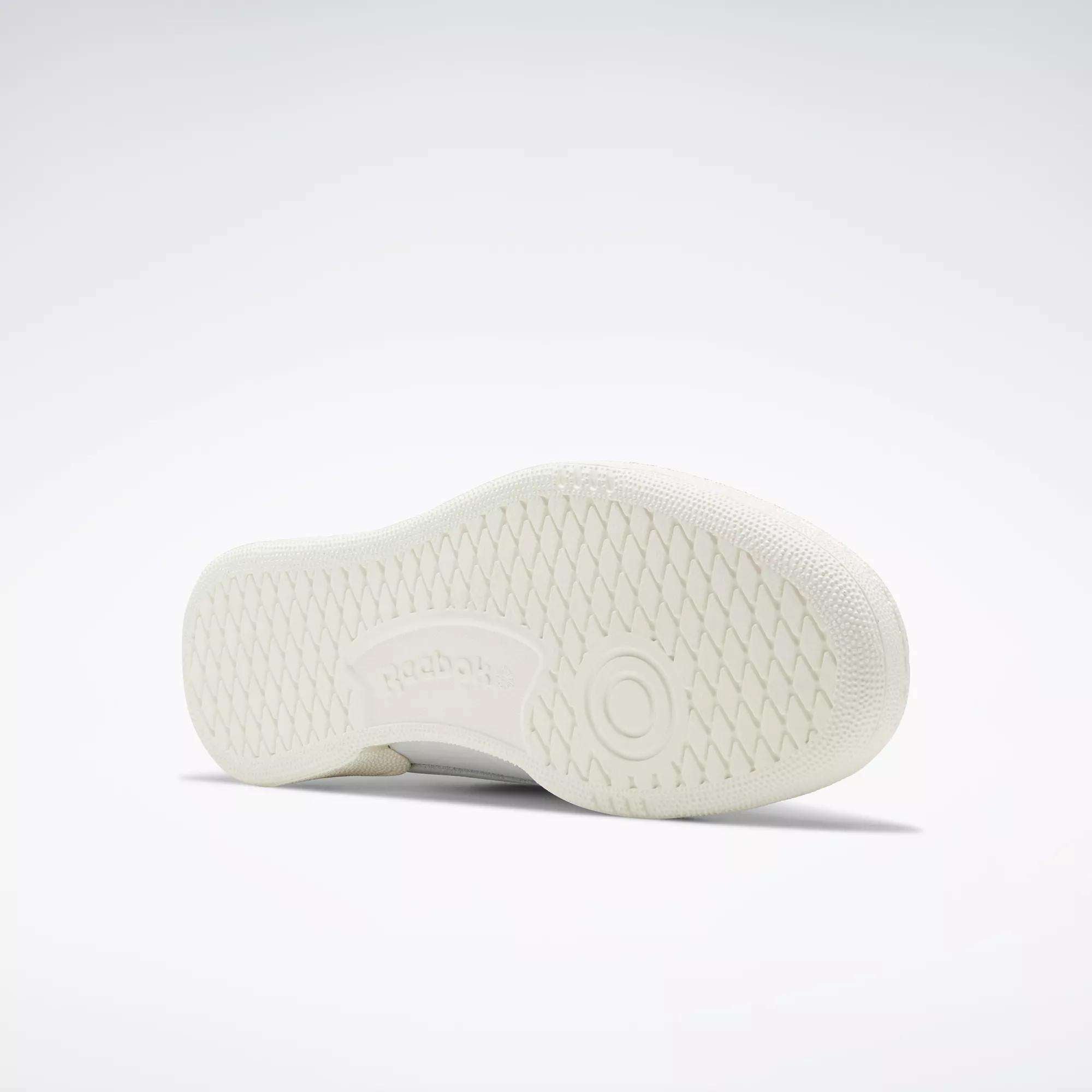 The Reebok Club C 85 Vintage Shoe in white is pictured on its side, revealing a textured sole with a circular pattern and an embossed Reebok logo, set against a plain light gray background.