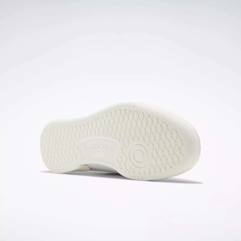 The Reebok Club C 85 Vintage Shoe in white is pictured on its side, revealing a textured sole with a circular pattern and an embossed Reebok logo, set against a plain light gray background.