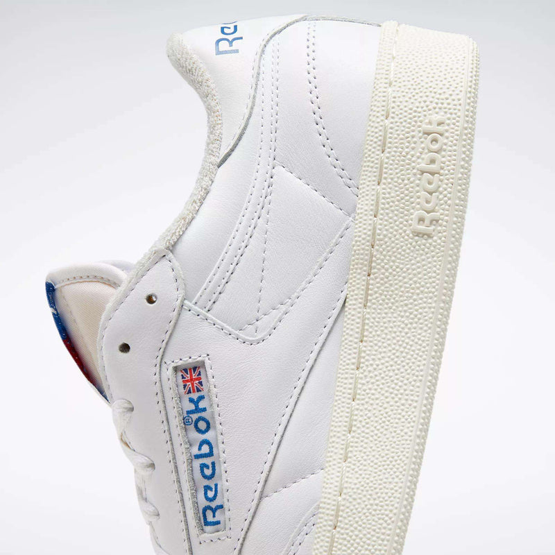 A close-up of Reebok Club C 85 Vintage Shoes shows their white leather upper, textured cream sole with lightweight cushioning, blue and red Reebok logo, and Union Jack flag on the side, angled to reveal both the side and part of the sole.