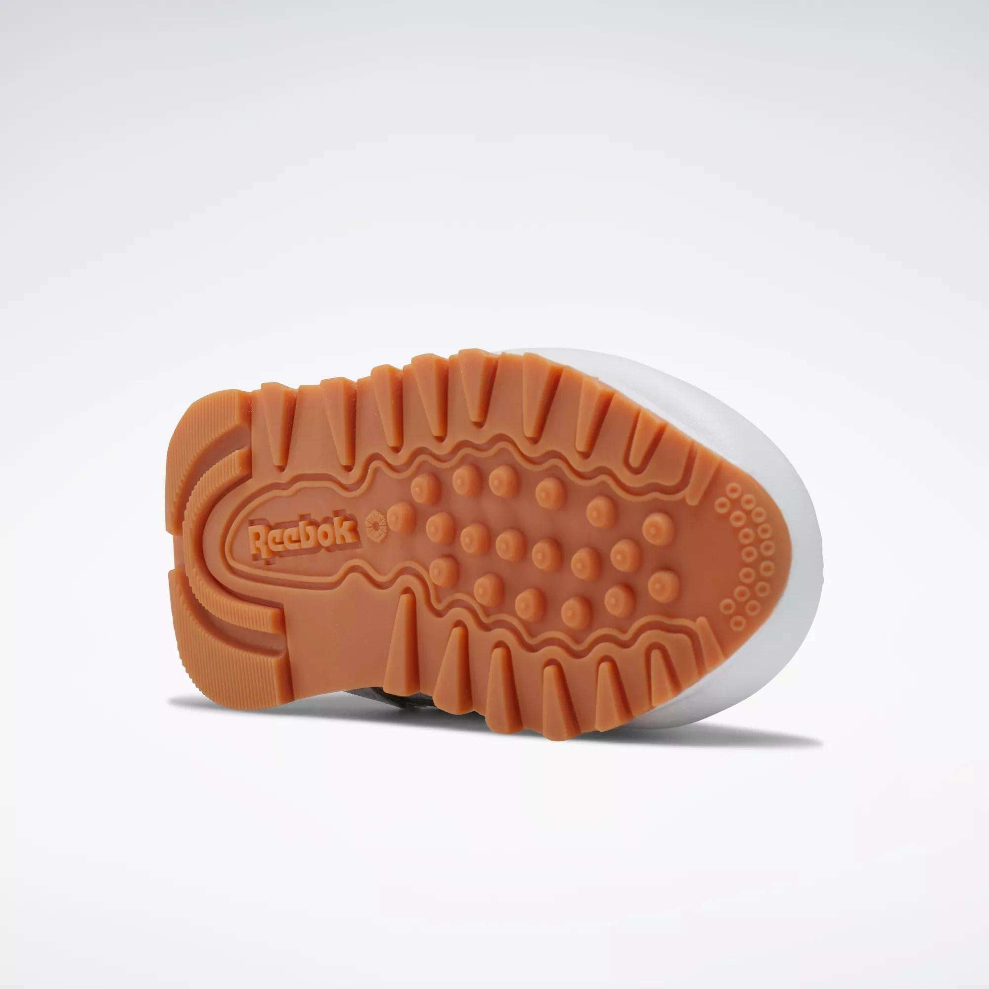 The image shows the sole of a Reebok Kids' Classic Leather Shoe for babies and toddlers, featuring a gum rubber outsole with textured tread and the Reebok logo near the heel. The shoe is displayed sole-up against a plain background.