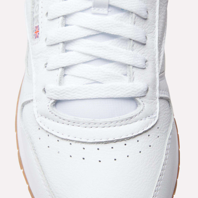 Close-up of the toe and laces of a white Reebok Classic Leather Shoes with perforations, a tan outsole, and a small Union Jack flag patch on the side.