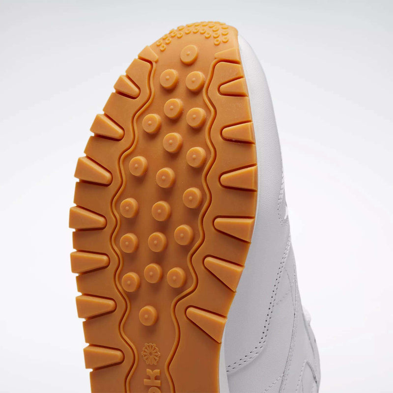 Close-up of the Reebok Classic Leather Shoes sole with textured light brown rubber tread in circular and wavy patterns. The white leather upper stands out against a subtle gradient background.