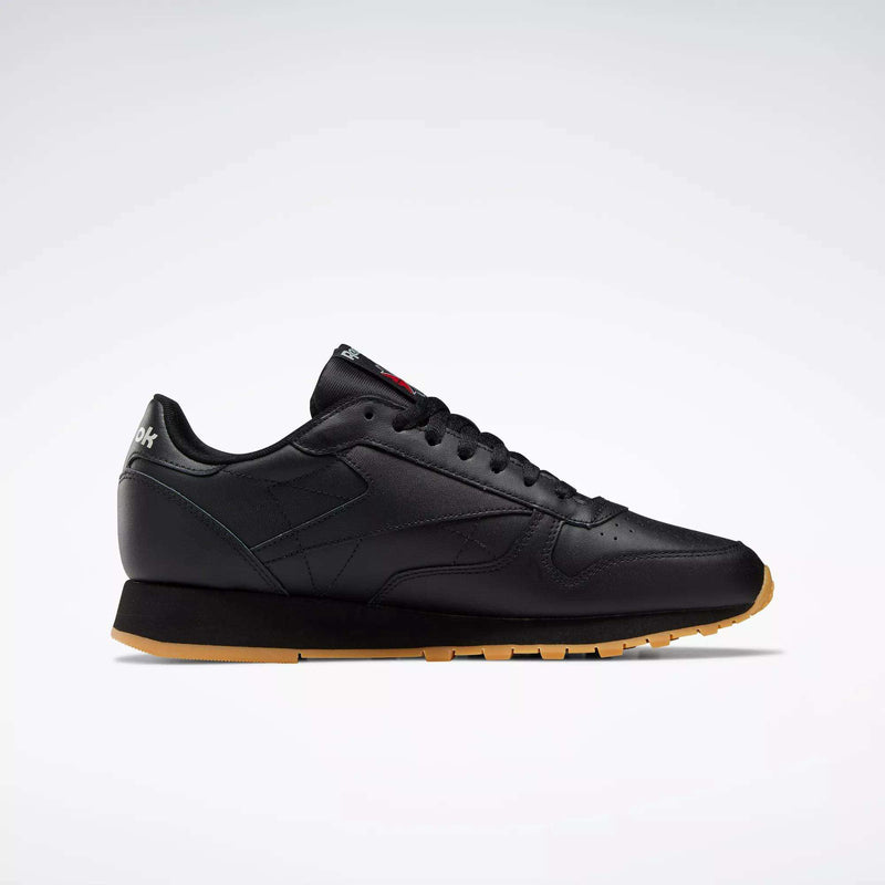 Side view of Reebok Classic Leather Shoes in black with a gum sole, black laces, minimal stitching, and low-cut design. These retro-inspired shoes have a durable leather upper and are shown on a plain light gray background.