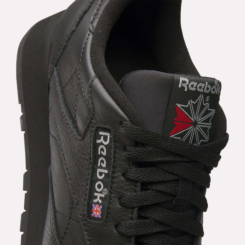 Close-up of a black Reebok Classic Leather Shoes sneaker, highlighting the tongue and side with stitched Reebok logos, red and white starburst graphic, and a small Union Jack. Durable leather upper delivers a retro-inspired Reebok style.