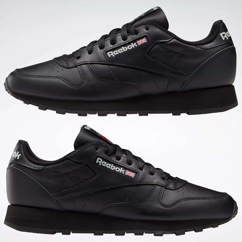 Two Reebok Classic Leather Shoes in black are displayed side by side on a white background, featuring the Reebok logo and British flag near the laces, minimalist stitching, and black rubber soles.