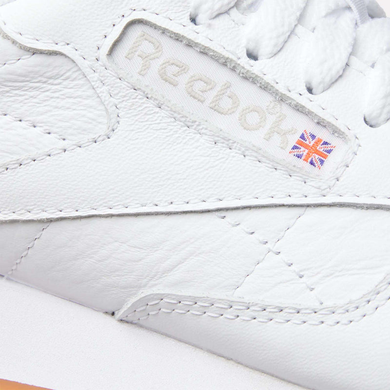 Close-up of Reebok Women's Classic Leather Shoes, featuring the embossed logo and a small Union Jack flag on the leather upper near the laces, detailed stitching, and a cushioned midsole.