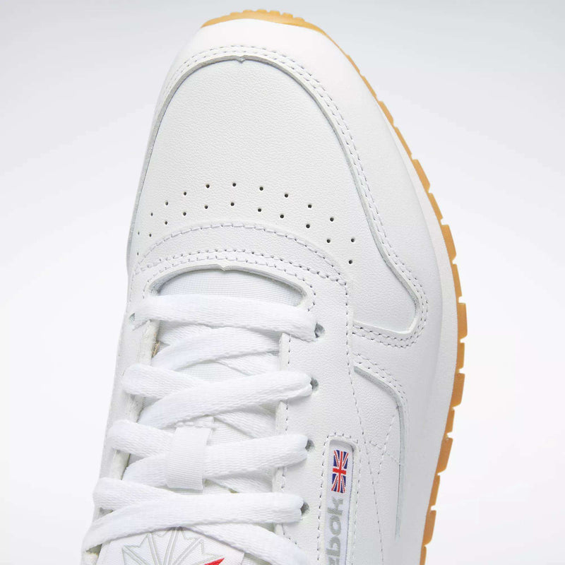 Close-up of Reebok Women's Classic Leather Shoes in white, featuring white laces, perforated toe, cushioned midsole, small British flag logo on the side, and a light brown rubber sole.