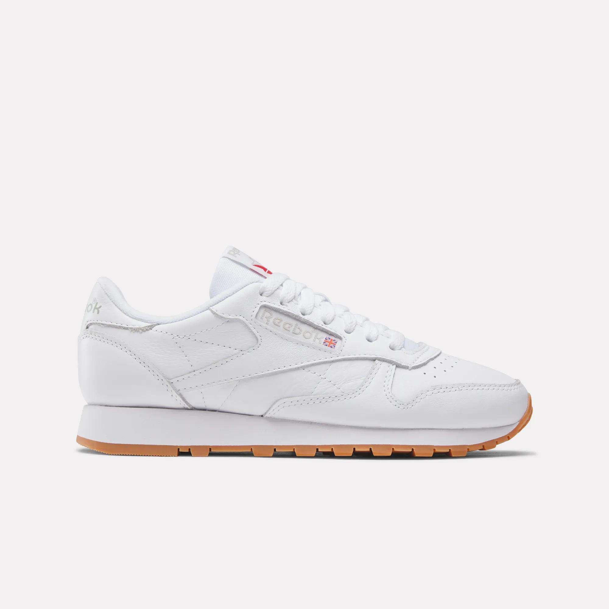 The Reebok Women's Classic Leather Shoes feature white leather, subtle branding, white laces, a cushioned midsole, and a light brown gum sole, shown in side profile on a plain white background.