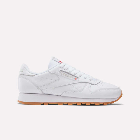 The Reebok Women's Classic Leather Shoes feature white leather, subtle branding, white laces, a cushioned midsole, and a light brown gum sole, shown in side profile on a plain white background.