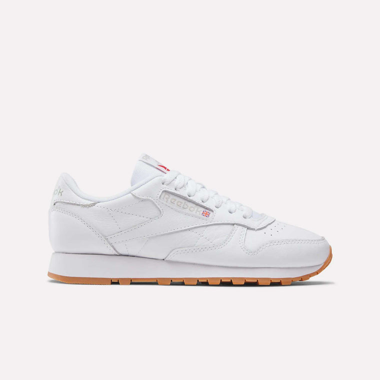The Reebok Women's Classic Leather Shoes feature white leather, subtle branding, white laces, a cushioned midsole, and a light brown gum sole, shown in side profile on a plain white background.