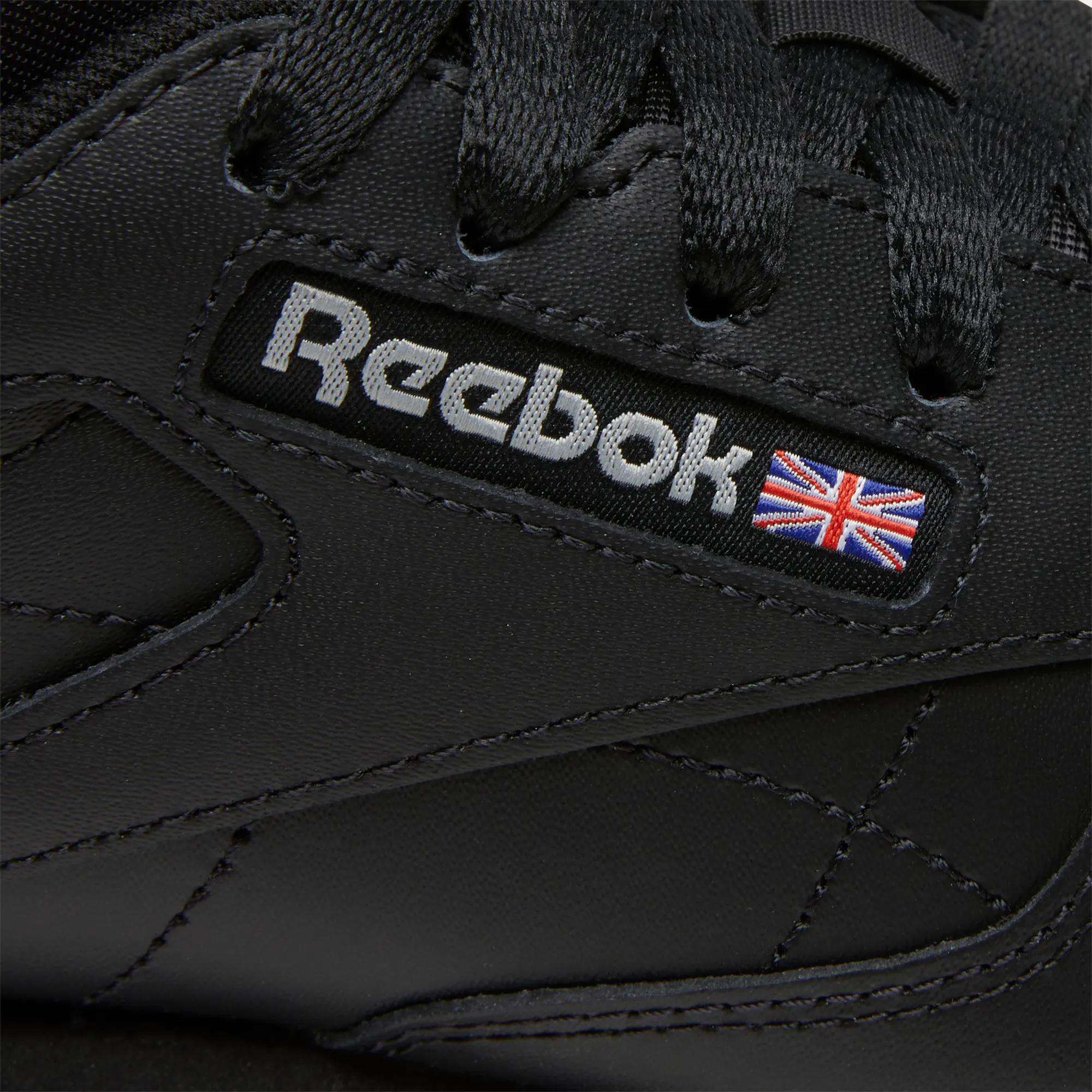 Close-up of Women's Classic Leather Shoes by Reebok, featuring the iconic logo, a stitched Union Jack flag on the side, black laces, visible leather stitching, and a cushioned midsole for comfort.
