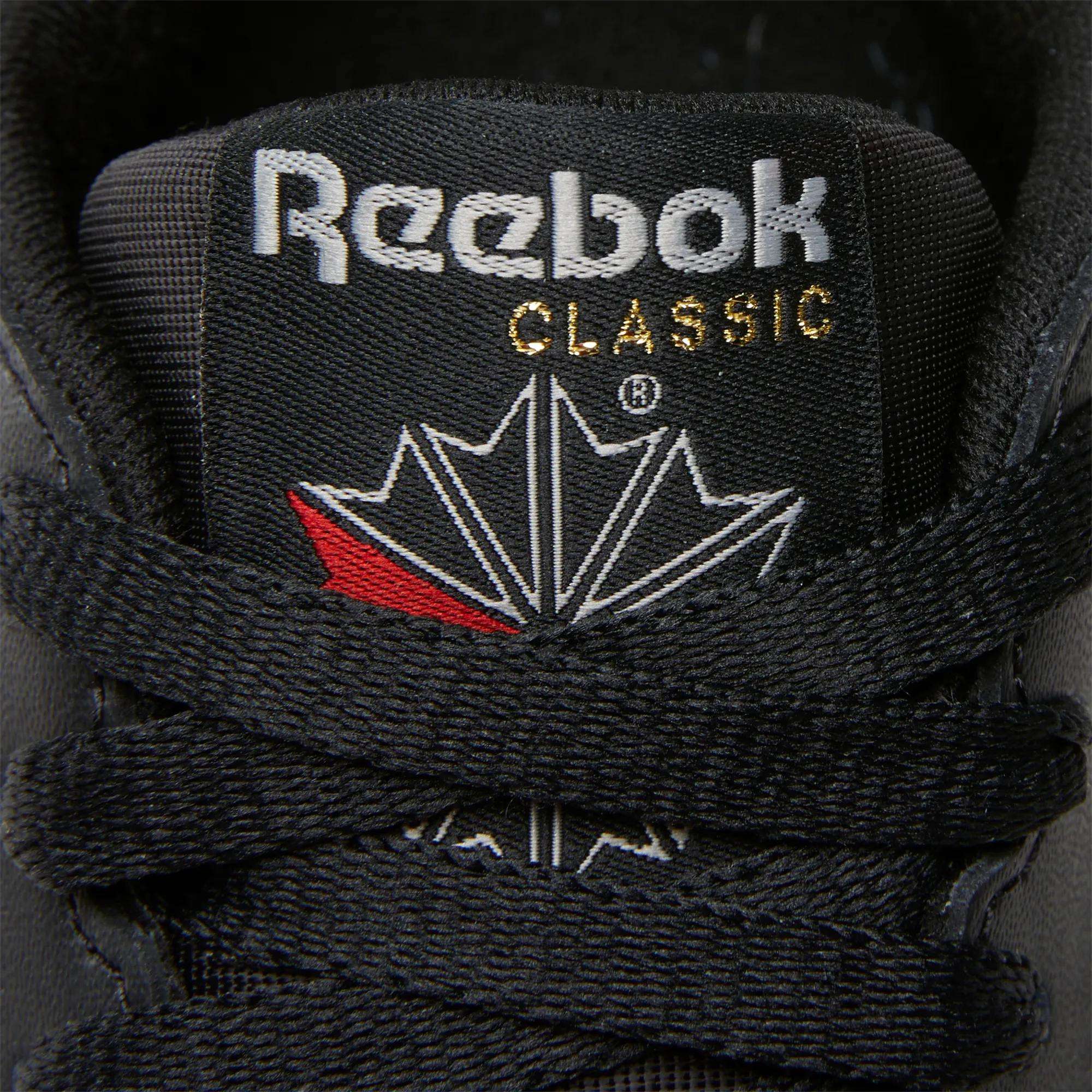Close-up of the black tongue on Reebok Women's Classic Leather Shoes with black laces, a white Reebok logo, gold "CLASSIC" text, red and white maple leaf design, and a cushioned midsole for comfort.