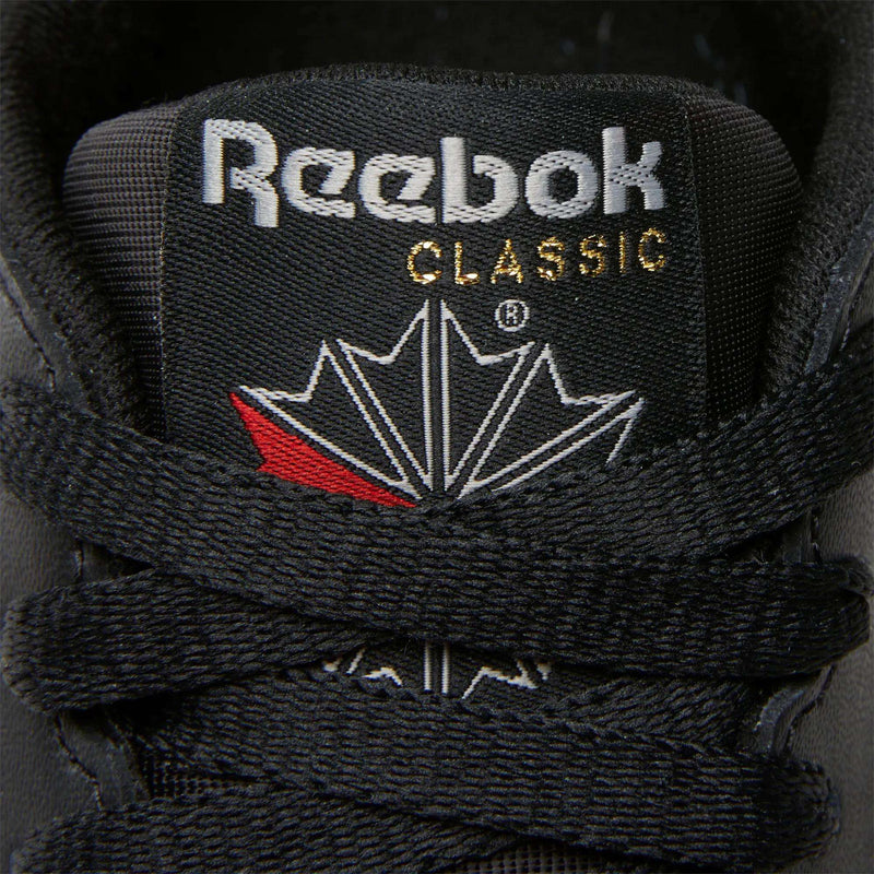 Close-up of the tongue on Reebok Women's Classic Leather Shoes, showing the Reebok logo, gold CLASSIC lettering, a red and white maple leaf design, and black laces—an iconic detail from the brand.