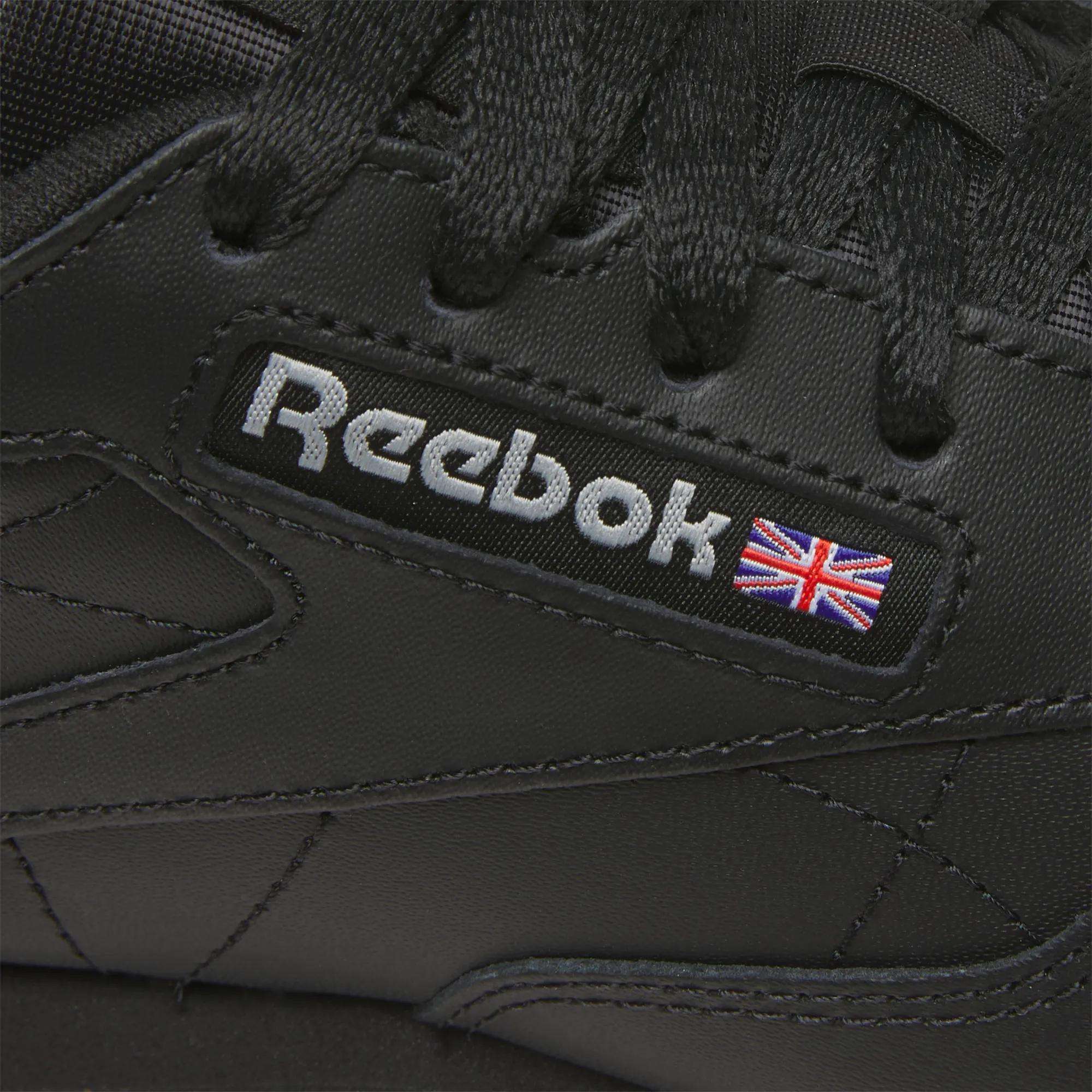 Close-up of Reebok Women's Classic Leather Shoes in black, featuring a stitched Reebok logo with a small Union Jack on the side, black laces, and a cushioned midsole for comfort.